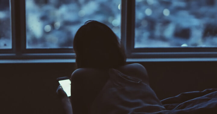 A photo of a woman lying on a bed while using her smartphone for the article "Studies: Health Effects of Blue Light Exposure"