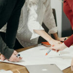 A mid-shot image of individuals standing up during a meeting for the article "Top-Down Management vs Bottom-Up Management: The Difference"