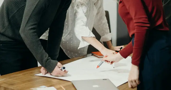 A mid-shot image of individuals standing up during a meeting for the article "Top-Down Management vs Bottom-Up Management: The Difference"