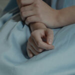 A close-up image of the hands of a patient sitting in a hospital bed for the article "Private Health Insurance as a Rent-Seeking Middleman"