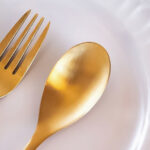 A tight shot of golden spoon and fork on an empty white plate for the article "Study: How Intermittent Fasting Slows Hair Growth"